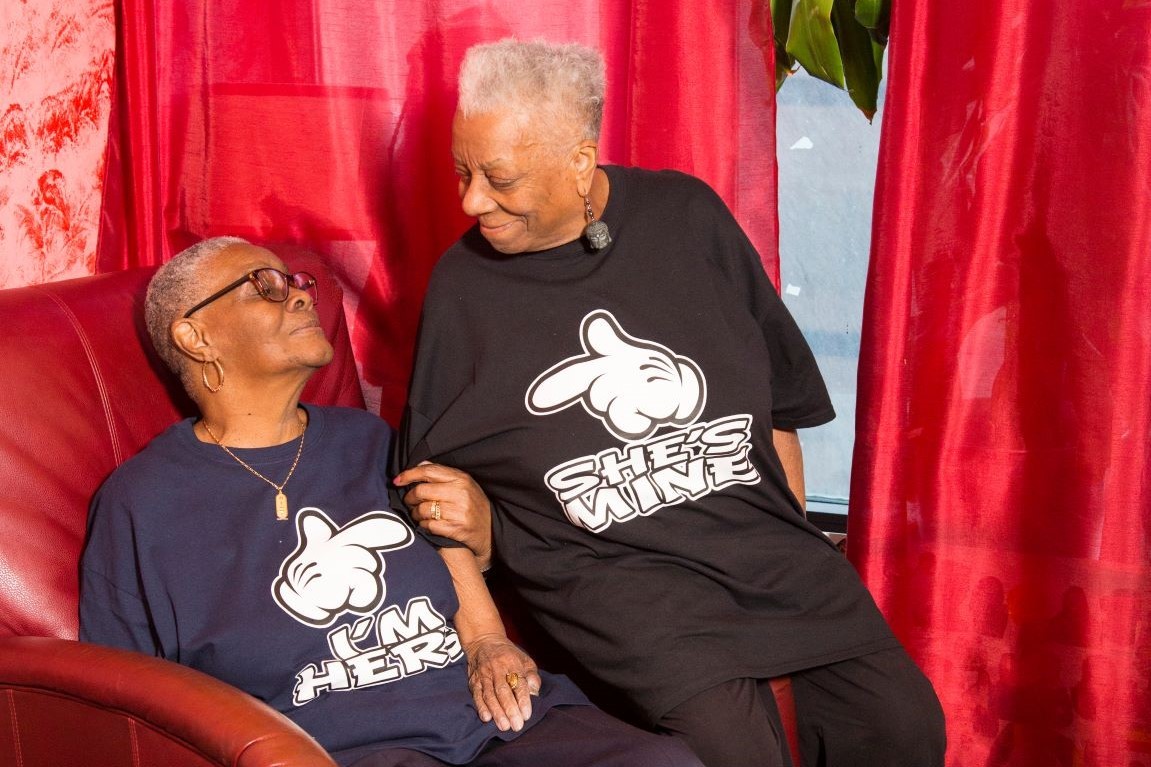 Two older adults sit together, wearing matching shirts; one shirt reads “I’M HERE” and the other reads “SHE’S MINE.” They smile and look at each other, seated by red curtains.