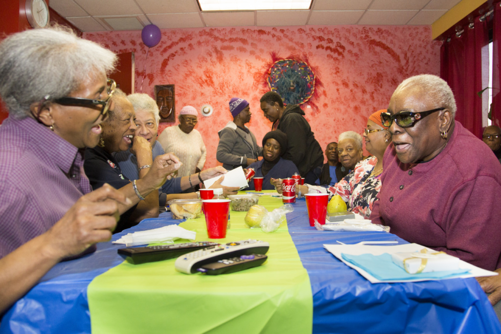 A group of seniors sit around a decorated table, talking and laughing, with food, drinks, and gifts on the table in a brightly colored room.