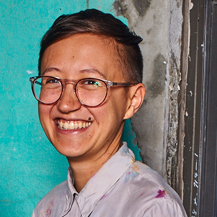 Person with short hair and glasses smiling, wearing a light-colored shirt, standing in front of a turquoise and gray textured wall.