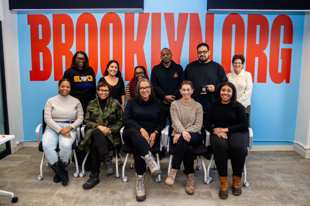 Twelve people pose for a group photo in an office with a large red “BROOKLYN.ORG” sign on the blue wall behind them.