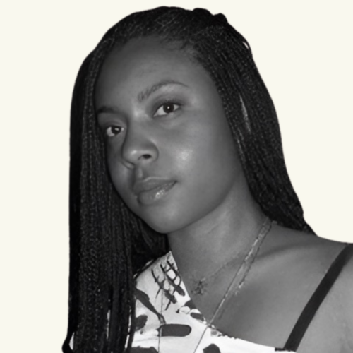 Black and white photo of a woman with long braided hair, looking at the camera, wearing a patterned top and layered necklaces.