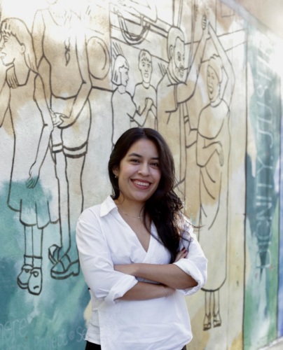 A woman with long dark hair stands smiling with arms crossed in front of a colorful mural depicting people engaged in various activities.