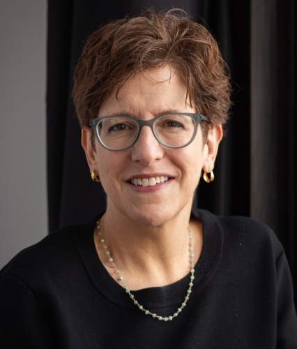 A person with short brown hair, glasses, and gold hoop earrings, wearing a black top and a beaded necklace, smiles at the camera in front of a dark background.