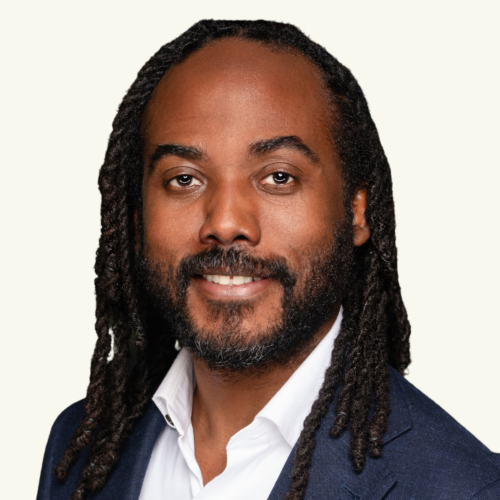 A man in a navy blazer and white shirt with long, dark, braided hair and a trimmed beard smiles at the camera against a light background.
