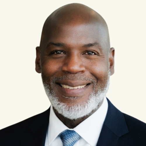 A man with a bald head and gray beard, wearing a dark suit, white shirt, and blue patterned tie, smiling at the camera against a plain light background.