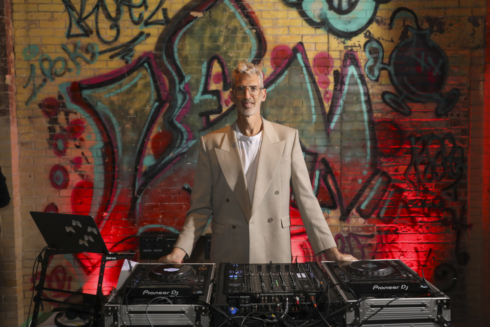 A person in a beige suit stands behind DJ equipment, with colorful graffiti and red lighting on a brick wall in the background.