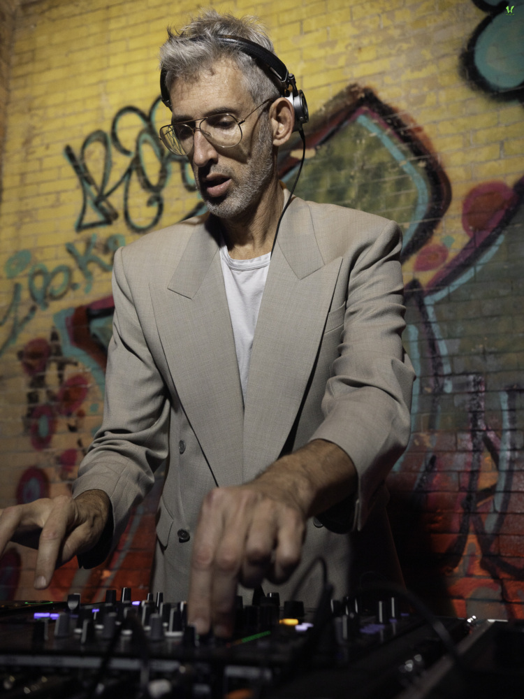 A man wearing headphones and a beige blazer operates a DJ mixer in front of a graffiti-covered brick wall.