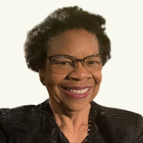 Smiling person with short curly hair, glasses, and a dark jacket, posed in front of a plain light background.