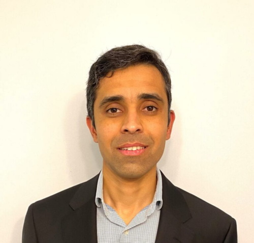 A man with short dark hair wearing a black blazer and light-patterned shirt stands against a plain white background, facing the camera.