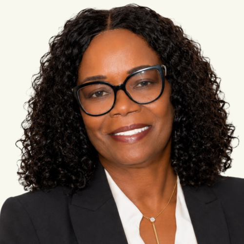 A woman with curly black hair, black glasses, and a black blazer over a white blouse smiles at the camera against a plain background.