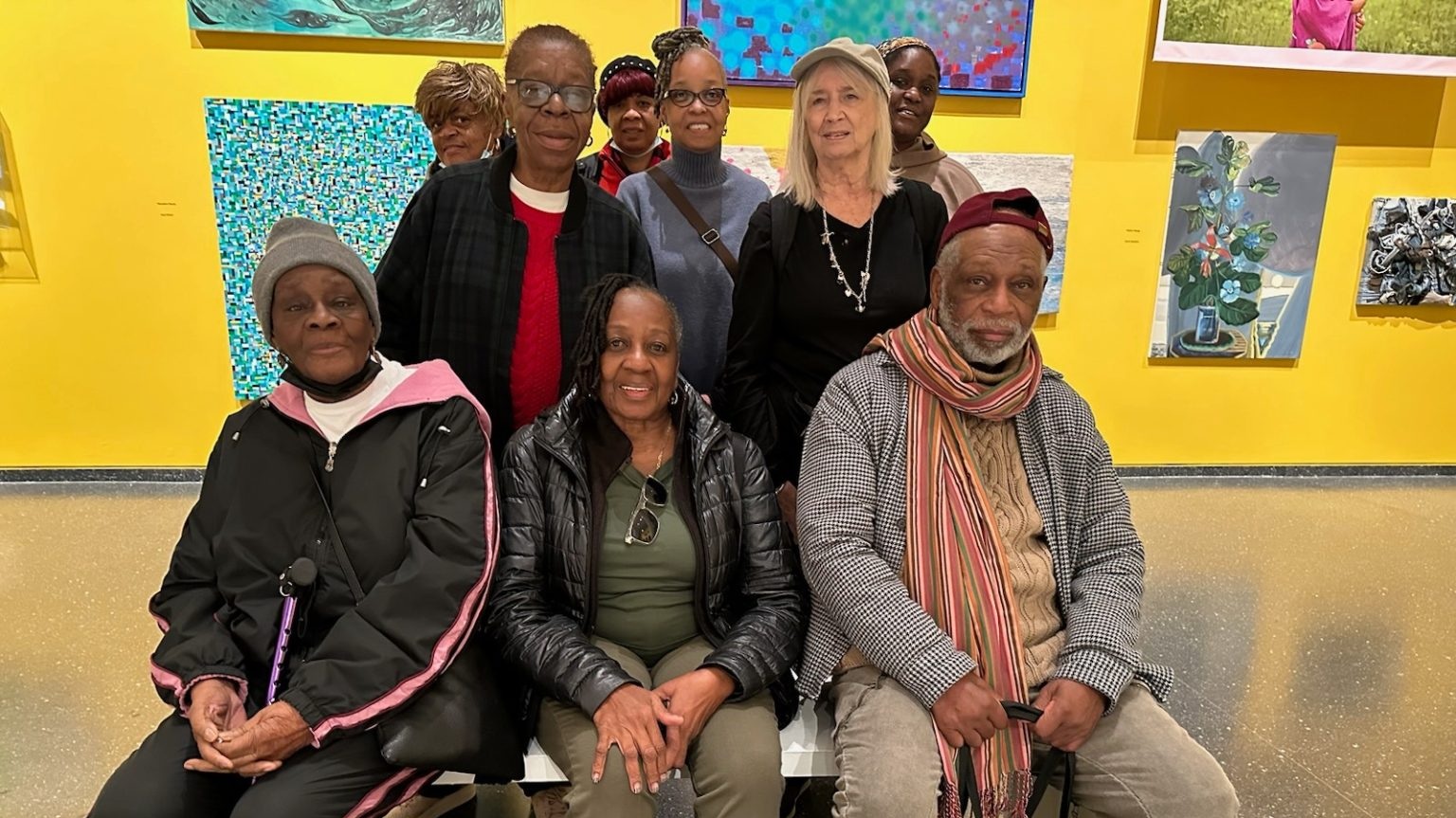 A group of nine adults pose for a photo on a bench in an art gallery with colorful paintings on a yellow wall behind them.