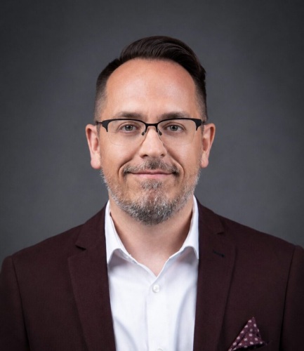 A man with short dark hair, glasses, and a trimmed beard wears a maroon suit jacket, white shirt, and polka dot pocket square, posing against a plain dark background.
