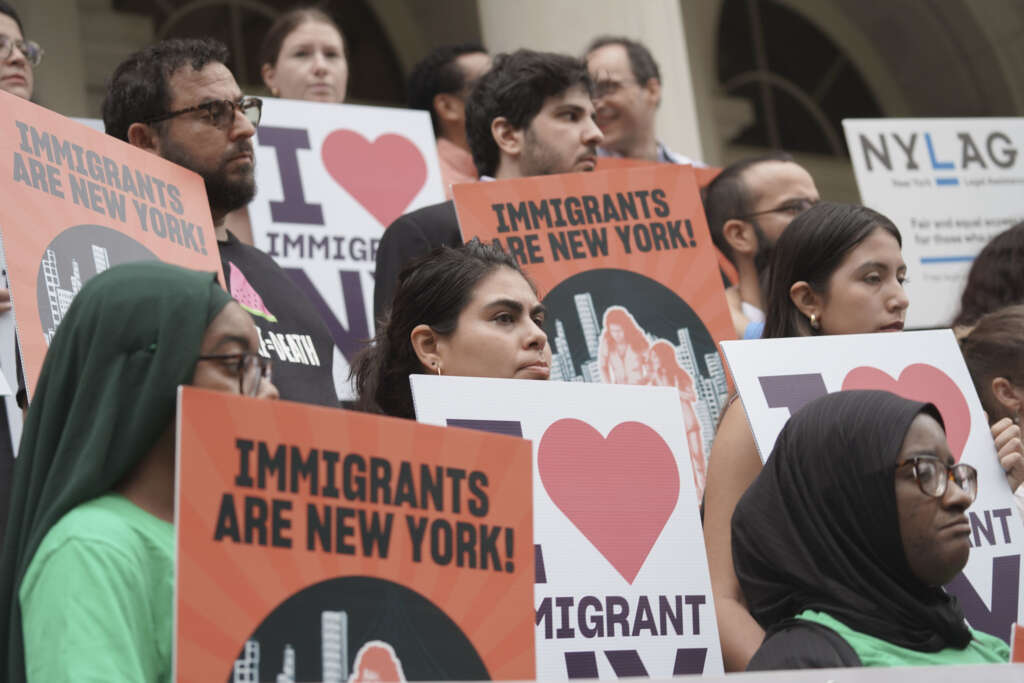 A group of people hold signs that say “IMMIGRANTS ARE NEW YORK!” and “I ❤️ IMMIGRANTS” at a public rally.