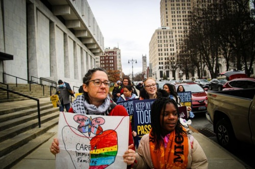 A group of people hold signs supporting immigrant families during a street protest in an urban area with tall buildings and parked cars nearby.