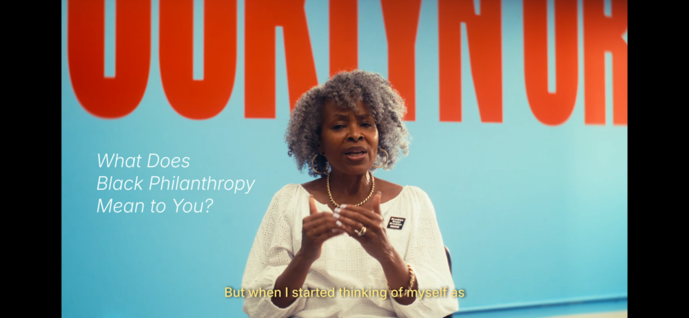 An older woman with gray curly hair speaks in front of a blue wall with bold red letters; text reads, “What Does Black Philanthropy Mean to You?” and yellow captions appear at the bottom.