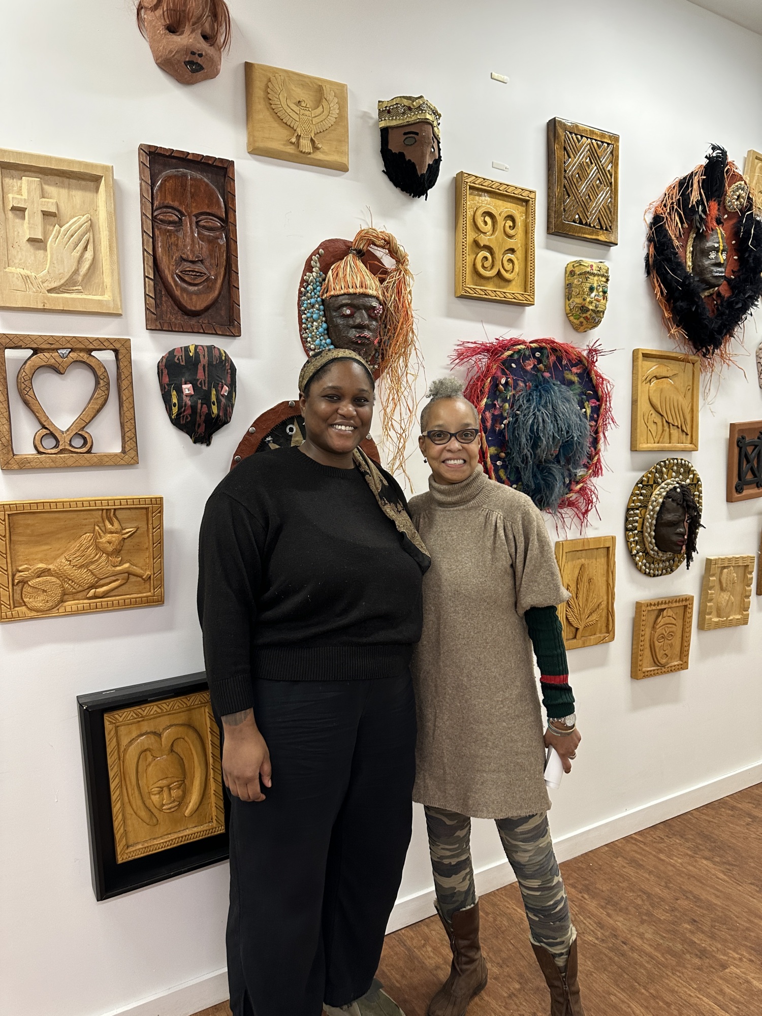 Two women stand smiling in front of a wall displaying various wooden and beaded masks and carvings.