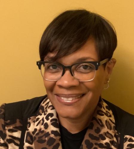 A person with short hair and glasses smiles at the camera, wearing a leopard print jacket in front of a yellow wall.