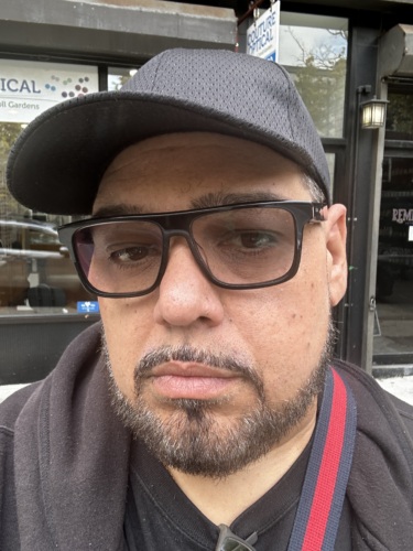 A man wearing glasses, a black cap, and a black jacket stands outdoors in front of a building with glass windows and signage.