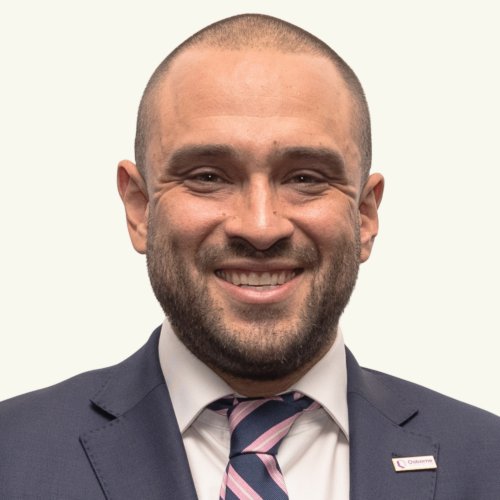 A man in a suit and striped tie is smiling at the camera, wearing a lapel pin on his left jacket collar.
