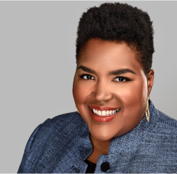 Smiling person with short hair wearing a blue textured blazer and gold earrings, posing in front of a plain gray background.