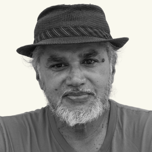 Gray-bearded man wearing a textured fedora and a T-shirt, looking directly at the camera with a neutral expression against a plain background.