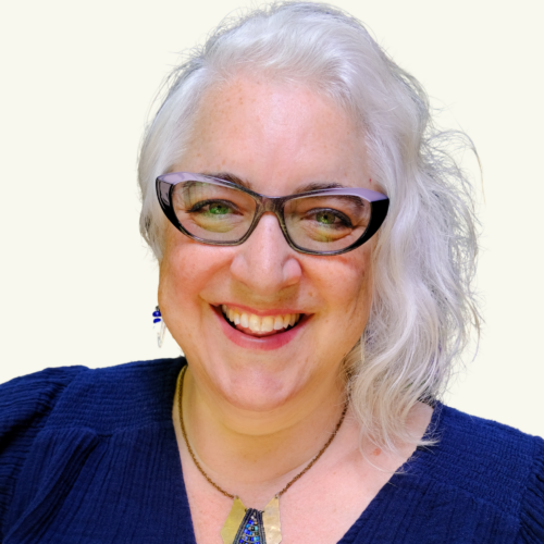 Smiling woman with white hair and glasses wearing a navy blue top, blue earrings, and a pendant necklace, facing the camera against a plain light background.