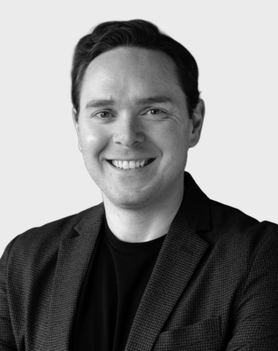 A man with short dark hair, wearing a textured blazer and a dark shirt, smiles while standing in front of a plain light background.