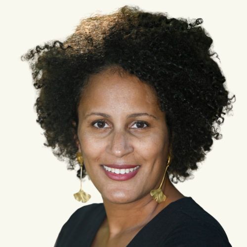 Smiling person with curly hair, gold leaf-shaped earrings, and a black top, shown against a plain light background.