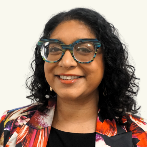 A person with curly dark hair, large blue and black glasses, and a colorful floral blazer is smiling at the camera against a plain background.