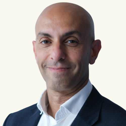 Bald man in a dark suit jacket and white shirt, smiling slightly, facing the camera against a plain light background.