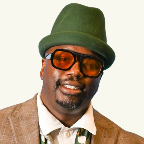 Man wearing an olive green hat, brown sunglasses, and a brown checked jacket over a collared shirt, smiling at the camera.