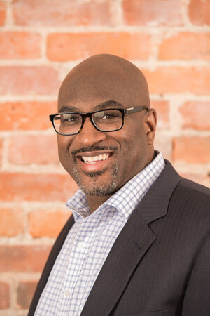 Smiling man with glasses in a suit jacket and checkered shirt stands in front of a brick wall.