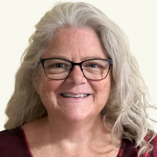 Smiling woman with long gray hair, wearing black glasses and a maroon top, pictured against a plain light background.
