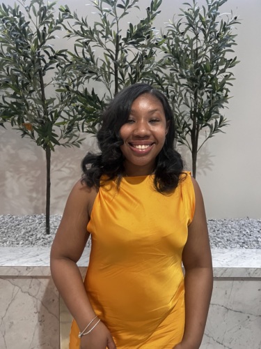 A woman in a sleeveless yellow dress stands indoors in front of two leafy green plants and a marble wall, smiling at the camera.