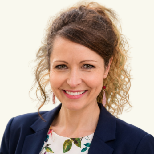 Woman with curly brown hair, wearing a navy blazer, floral top, and dangling earrings, smiling at the camera against a plain light background.