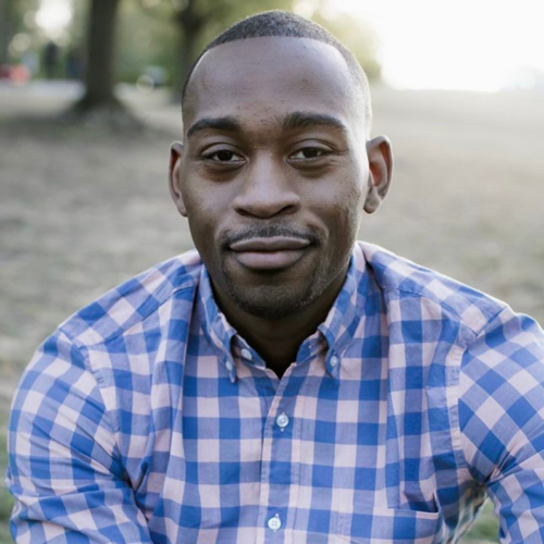 Man wearing a blue and pink checkered shirt sits outdoors on grass, looking directly at the camera with a slight smile.