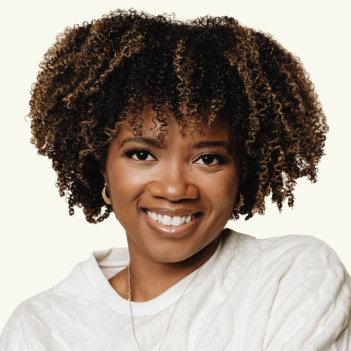 Woman with short curly hair and a white textured sweater, smiling at the camera against a plain light background.