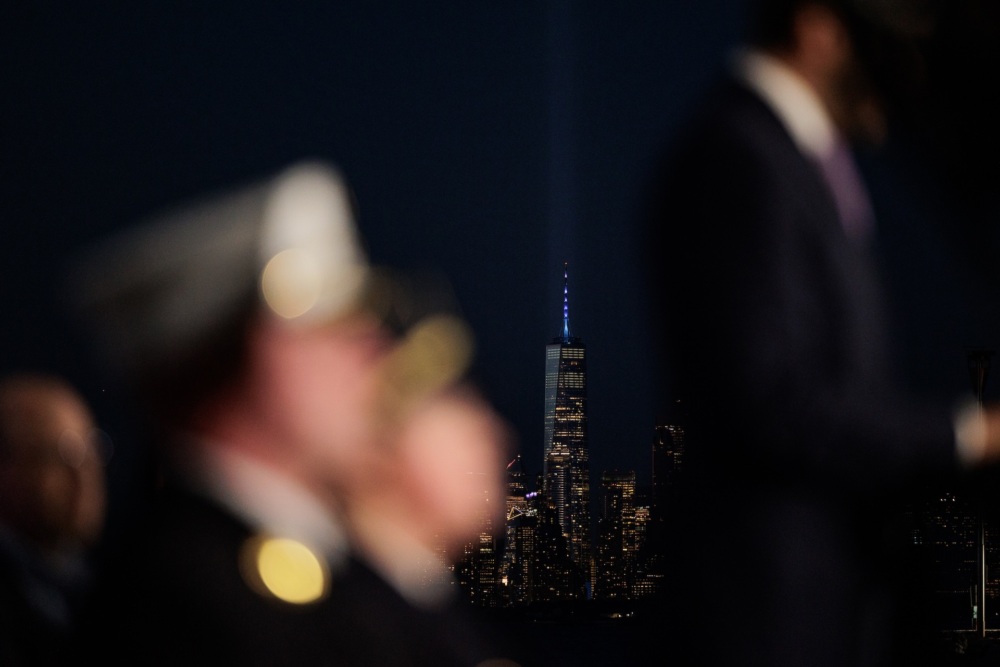 A blurred group of people in uniform in the foreground with the lit-up One World Trade Center and city skyline at night in the background.