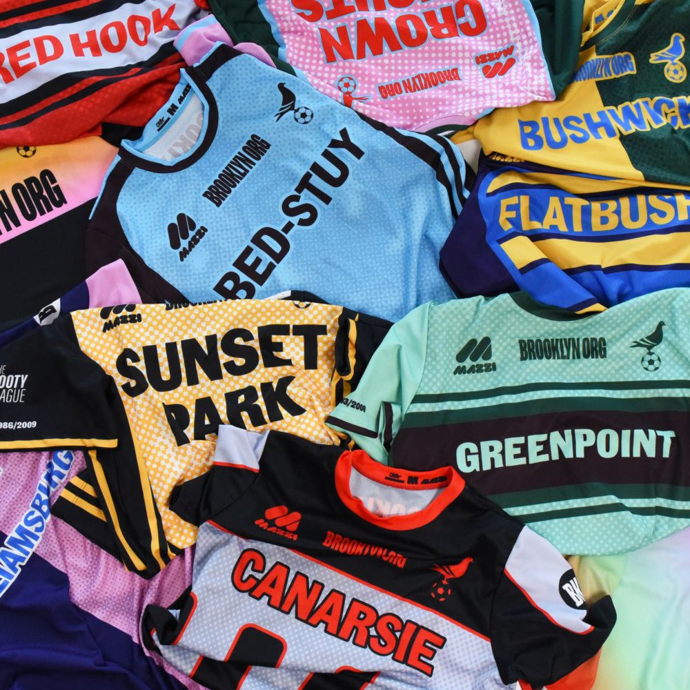 A variety of colorful Brooklyn sports jerseys are spread out, each labeled with neighborhood names like Bed-Stuy, Sunset Park, Greenpoint, Canarsie, Red Hook, and Bushwick.