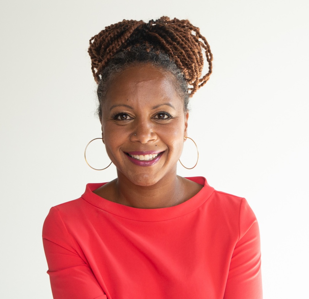 Woman with braided hair in a bun, wearing large hoop earrings and a bright red top, smiles at the camera against a plain white background.