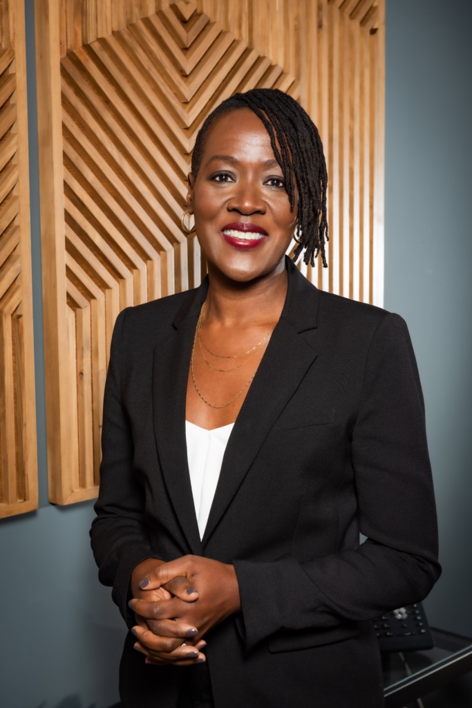 A person in a black blazer and white top stands indoors, smiling at the camera with hands clasped; wooden geometric panels hang on the wall behind them.