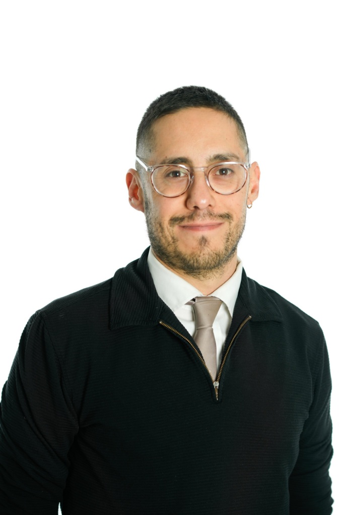 A person with short hair, glasses, and facial hair wearing a black top, white shirt, and beige tie stands against a white background.