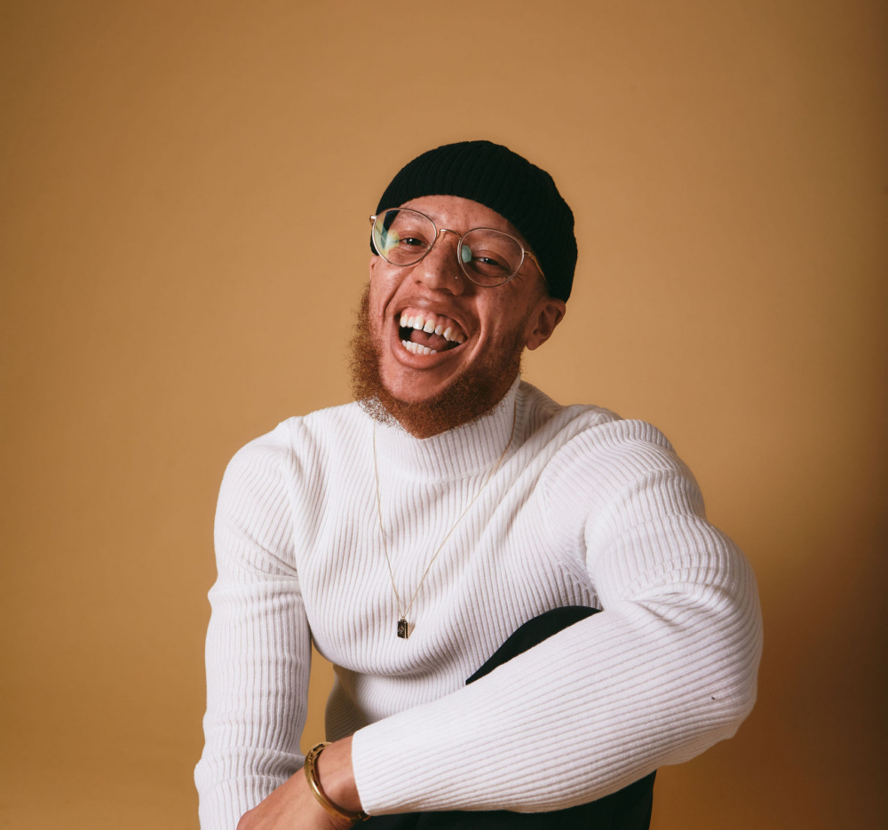 A man wearing a white sweater, black pants, glasses, and a black beanie sits on the floor against a beige background, smiling broadly.