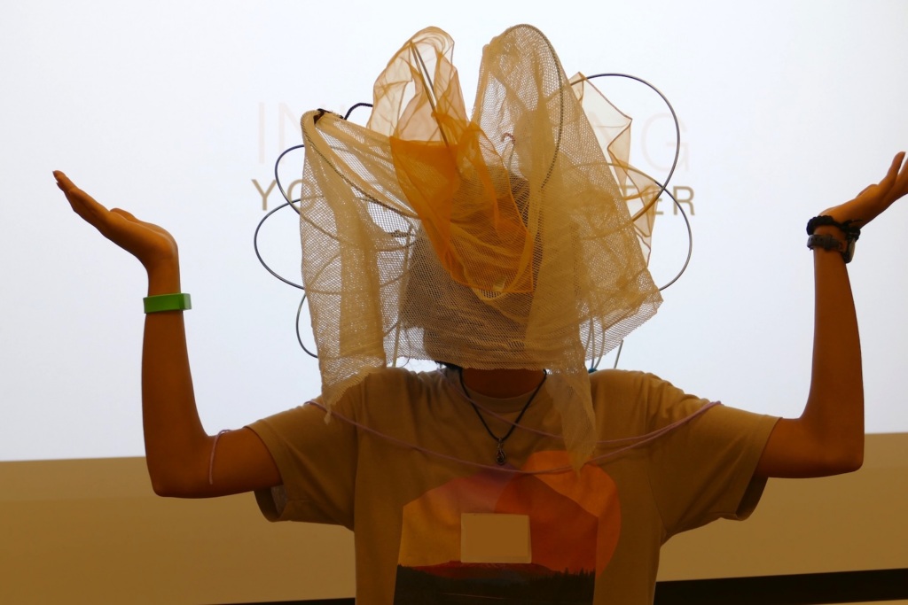 A person with arms outstretched wears a sculptural headpiece made of wire circles and sheer, layered netting.