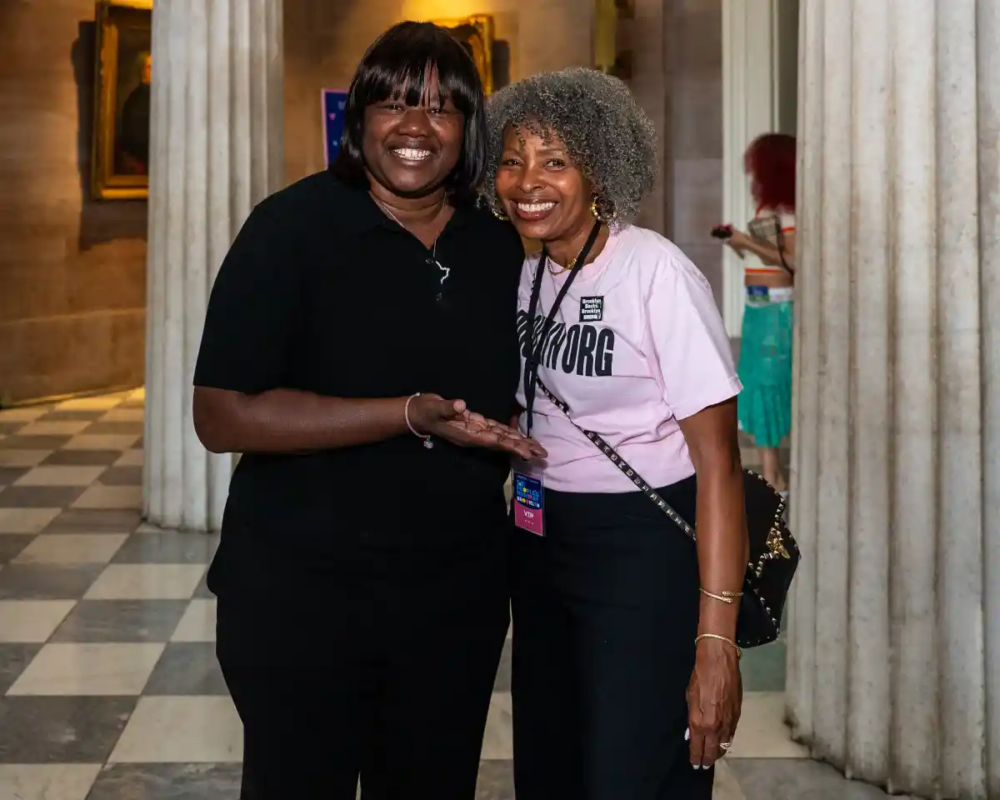 Two women stand together indoors, smiling at the camera. One wears a black shirt, the other a light pink shirt. They are surrounded by columned architecture and checkered flooring.