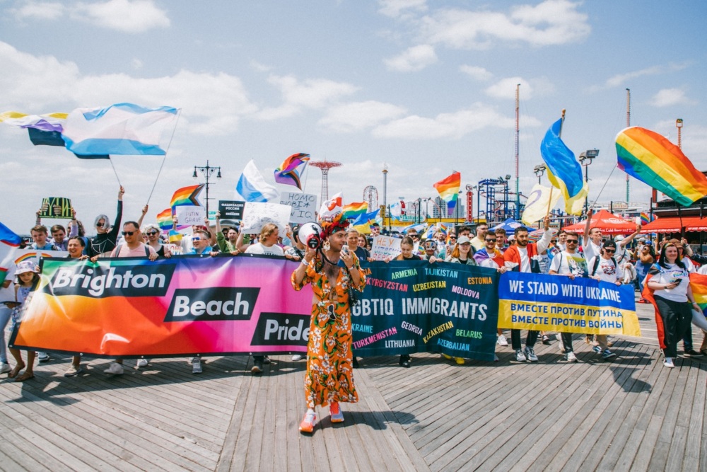 A large group of people marches on a wooden boardwalk at a Pride parade, carrying flags and banners that read “Brighton Beach Pride,” “LGBTQ IMMIGRANTS,” and “WE STAND WITH UKRAINE.”