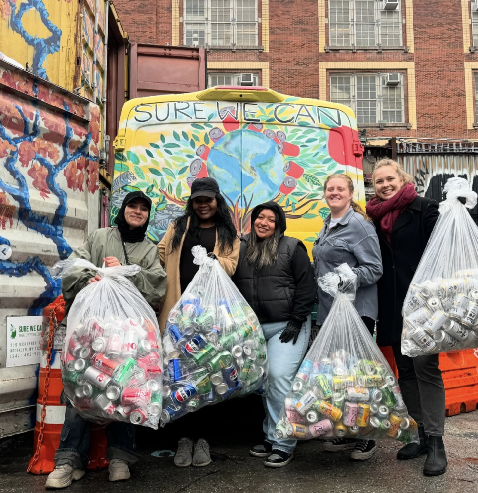 Five people stand in front of a painted van, each holding a large bag filled with aluminum cans, outside a brick building.