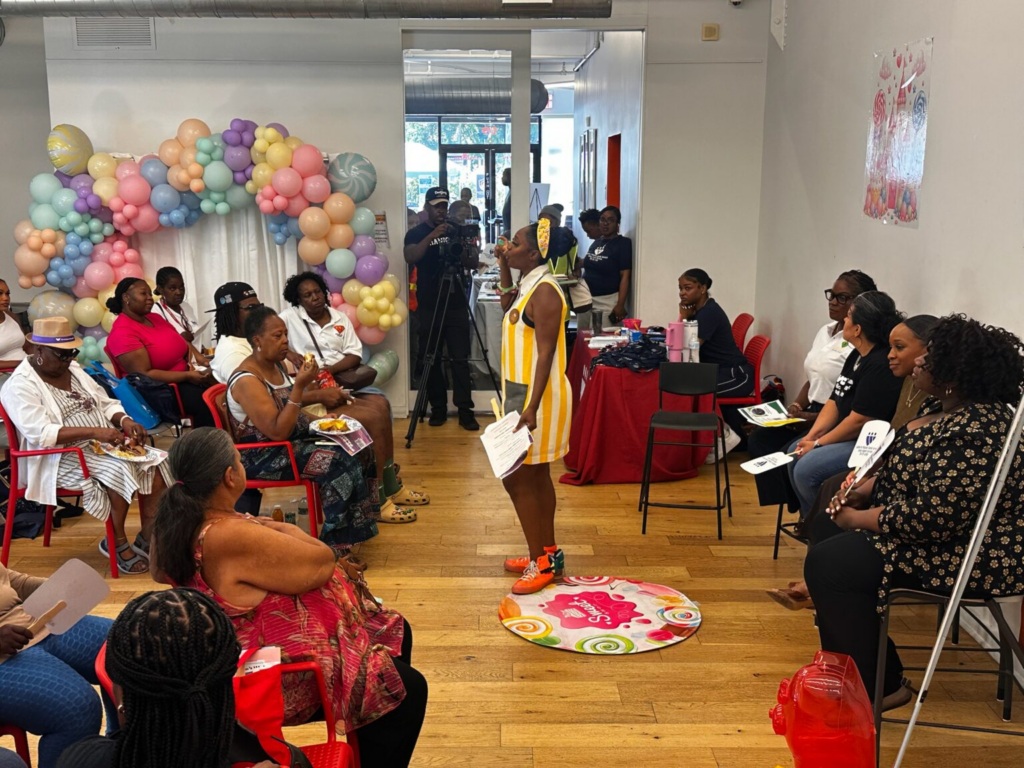 A person in a yellow and white striped dress stands in the center of a room presenting to a seated audience, with a balloon arch in the background.