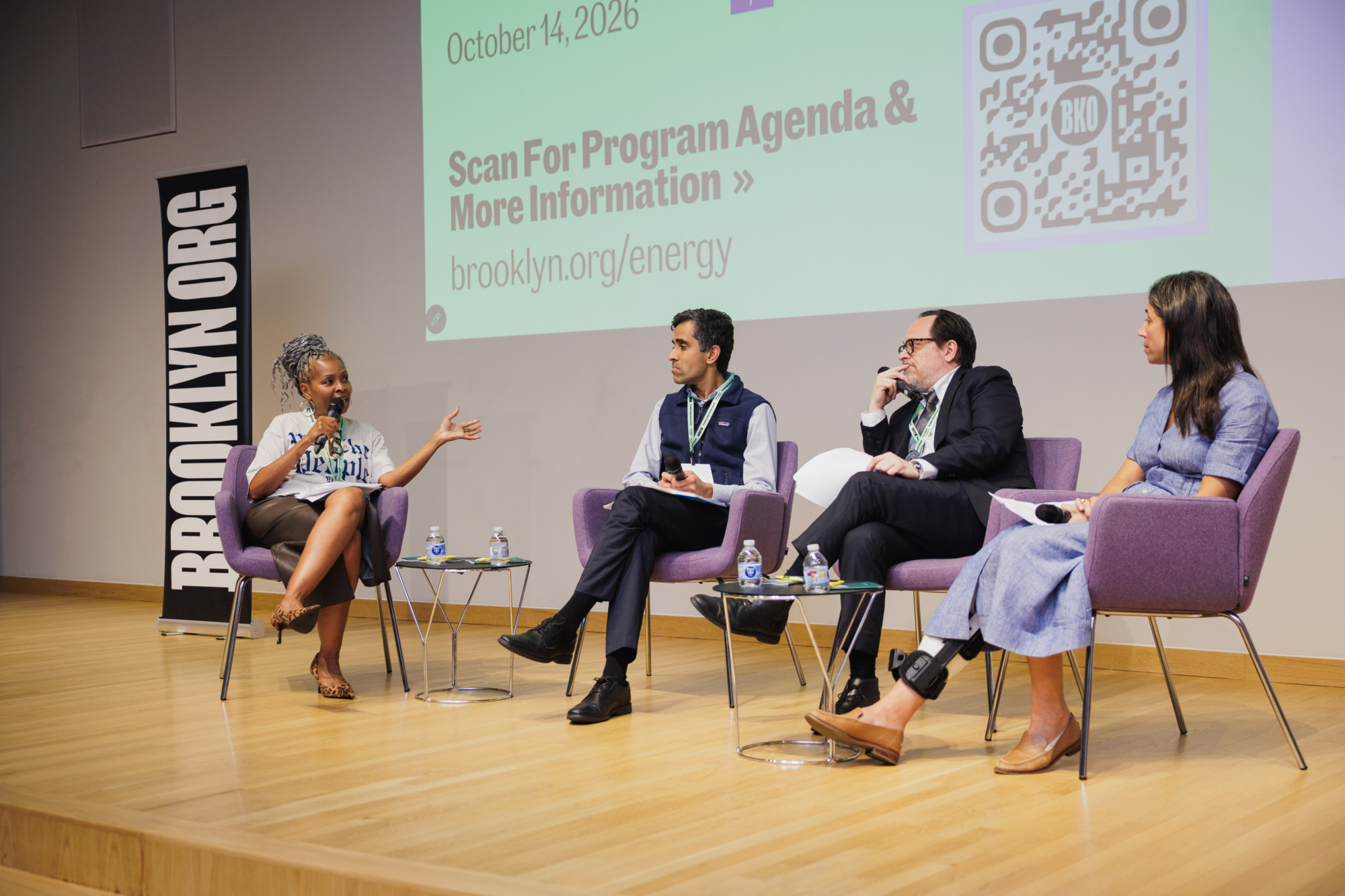 Four people sit in chairs on a stage during a panel discussion, with a large screen behind them displaying a QR code and event information for Brooklyn Org.