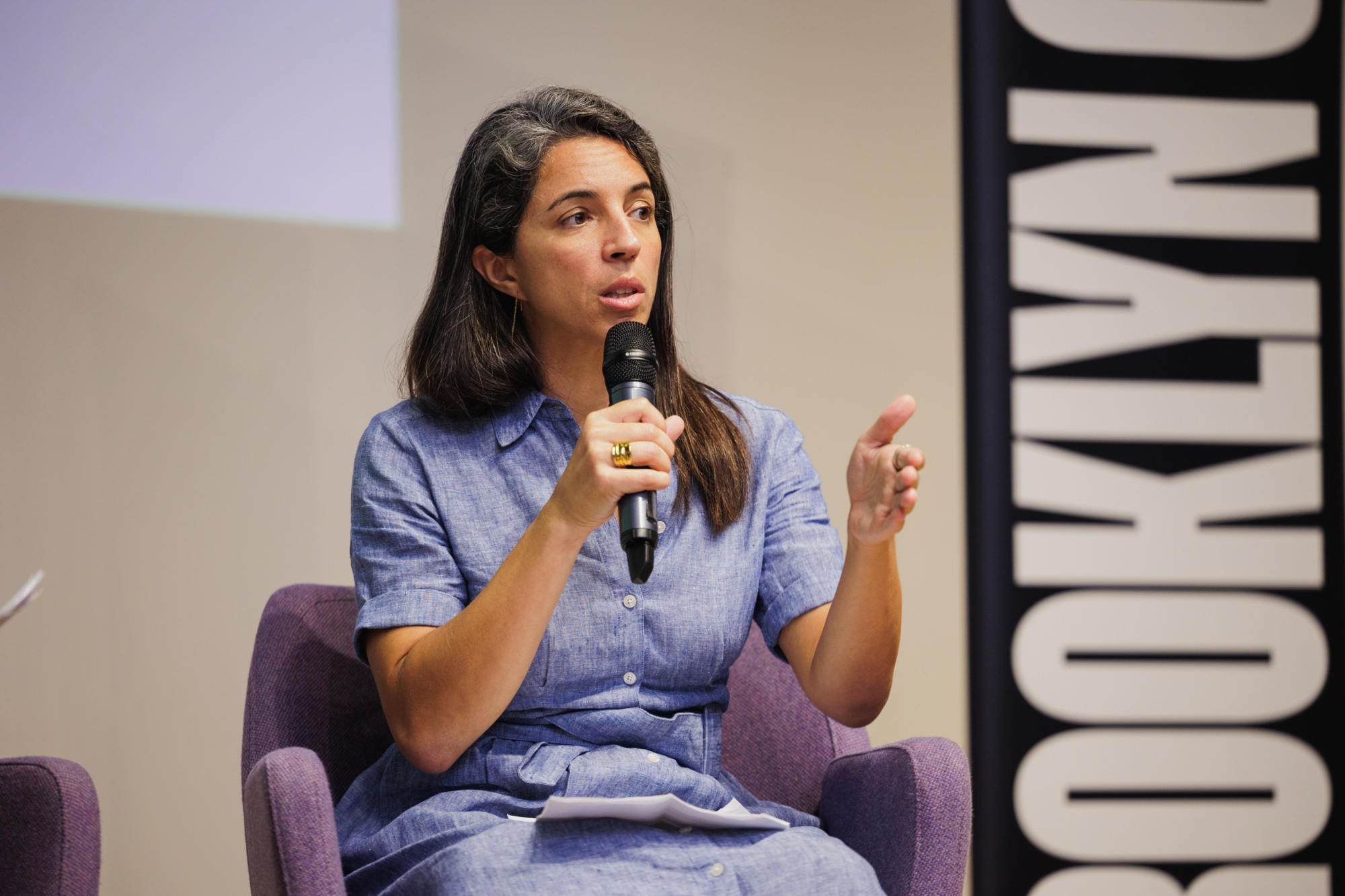 A woman in a blue dress sits in a chair, speaking into a microphone during a presentation.
